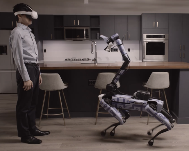 A person wears a virtual reality headset while facing a quadruped robot in the kitchen of an apartment.